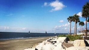 En strand med stenet kystlinje, palmer og sejlbåde i det fjerne.