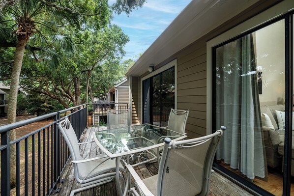 A balcony with a glass table and chairs, overlooking a garden with trees and a house in the background. - Lovely two Bedroom Villa! Open Kitchen! Short Ride to Beaches! Amenity access available for purchase (Seabrook Island)