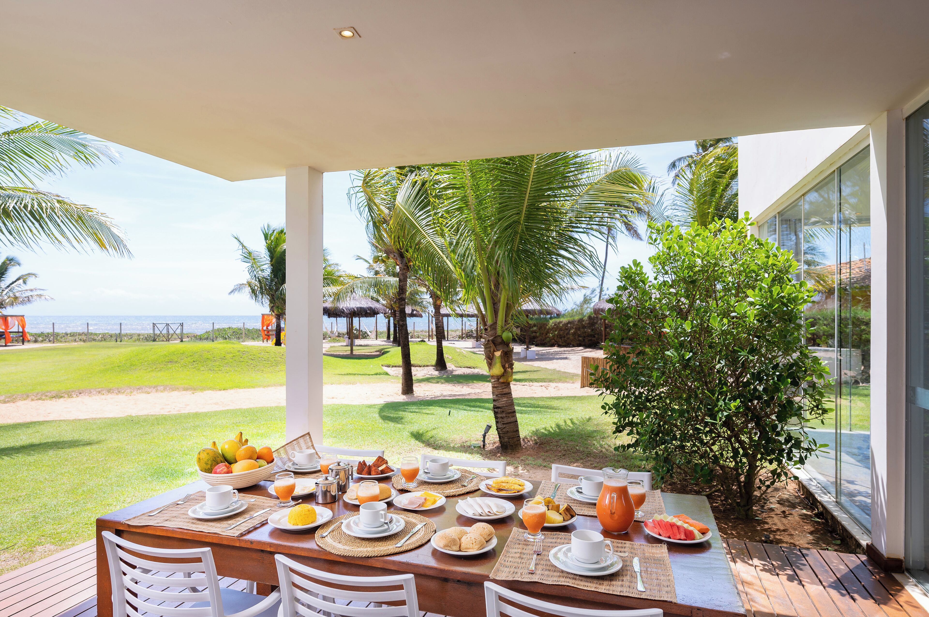 Mesa de desayuno dispuesta con platos, tazas y frutero, con vista a un área verde con palmeras y el océano.
