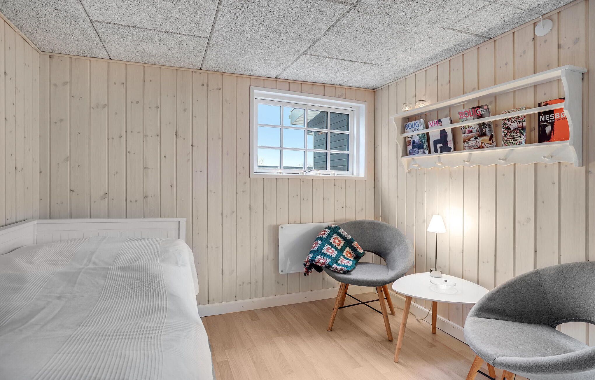 A room with wooden walls, a bed, a small table with a lamp, and a chair with a blanket.