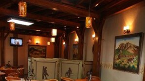Um restaurante com mesas e cadeiras de madeira redondas, uma televisão na parede e um quadro de montanhas.
