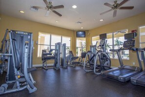 Un gimnasio con diferentes máquinas de ejercicio, ventiladores de techo y ventanas amplias.
