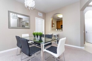 A dining room with a glass table, black and white chairs, a chandelier, and a kitchen visible through an archway.