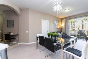Un espace repas moderne avec une table en verre et des chaises noires, un lustre et une vue sur un balcon aménagé avec du mobilier d’extérieur.