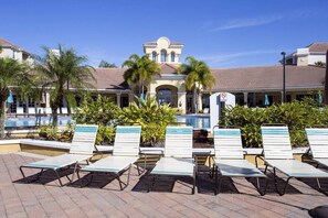 Un espace aménagé au bord de la piscine, avec des chaises longues, des palmiers et un bâtiment doté d’un toit en tuiles.