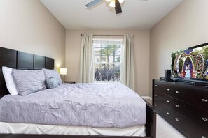 Une chambre à coucher avec un lit, un ventilateur de plafond, une fenêtre avec des rideaux et un téléviseur posé sur une commode.
