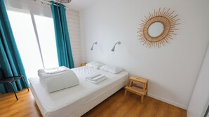 A bedroom with a bed, a wooden nightstand, a wall-mounted mirror, and a window with curtains.