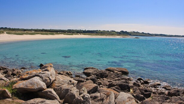 A rocky shoreline with clear blue water and a sandy beach in the background.