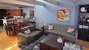 Een ruime woonkamer met een grijze bank, een houten eettafel en stoelen. In de achtergrond is een keuken zichtbaar met houten kastjes en een magnetron.