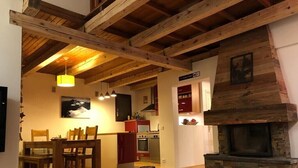 A cozy living room with wooden floors and a stone fireplace. There is a dining area with wooden chairs and a table. The kitchen is visible in the background with red cabinets and a stove. - Beau Chalet 9 persons, 5 rooms, 120m2 in Allos (Allos)