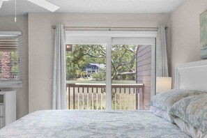 Un dormitorio con cama, ventana con persianas, vista a una casa y balcón con barandilla.