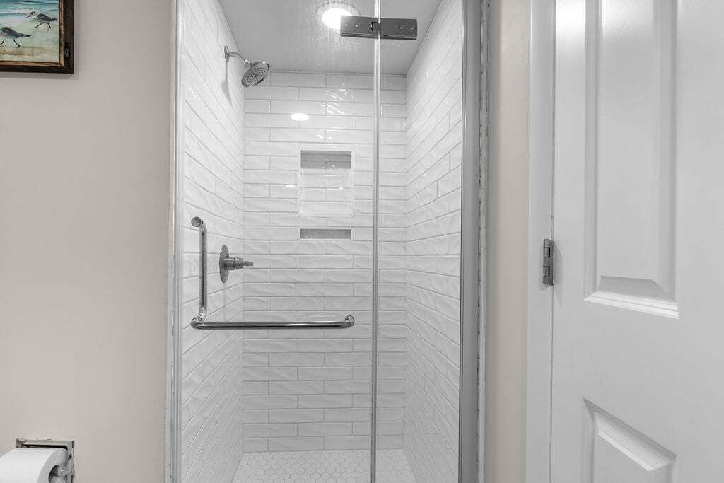 A glass-enclosed shower with white tiled walls, a metal grab bar, and a door with a white frame.