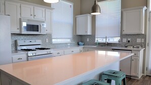 Une cuisine avec des meubles blancs, un grand îlot central, un four à micro-ondes, une cuisinière et deux tabourets.