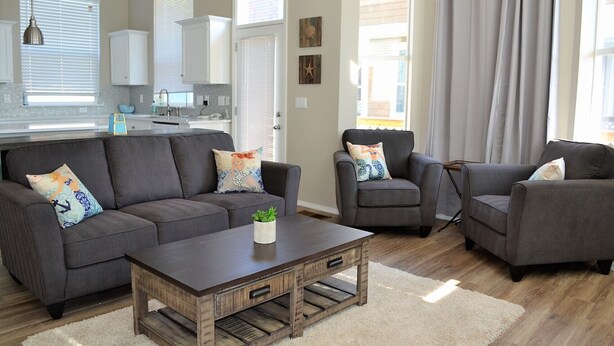 Un salon moderne avec un canapé gris, deux fauteuils, une table basse et une cuisine en arrière-plan.