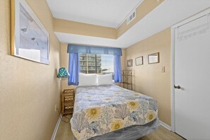 Quarto com cama, janela com cortinas azuis, mesa de cabeceira e porta.