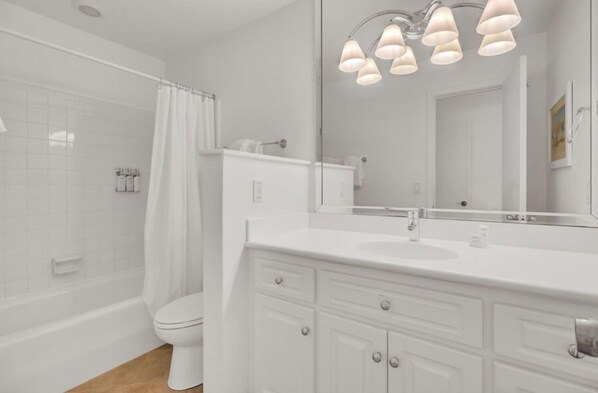 A modern bathroom with a white sink, vanity, and toilet. It features a large mirror, a shower with a curtain, and a ceiling light fixture.
