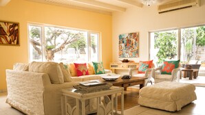 Un salón luminoso con un sofá, sillones y una mesa de centro. Hay ventanas amplias con vistas a árboles y un cuadro en la pared.