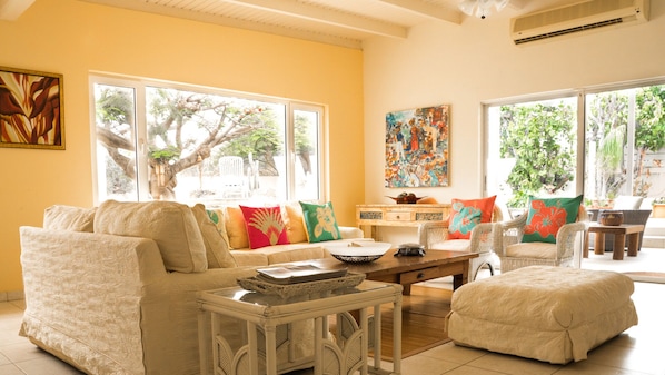 A bright living room with a sofa, armchairs, and a coffee table. There are large windows with a view of trees and a painting on the wall.