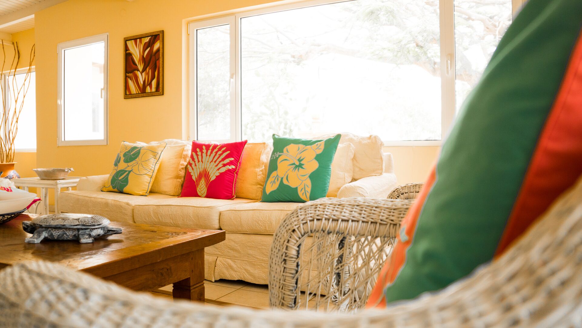 A cozy living room with a sofa, a wooden coffee table, and a wicker chair. There are decorative pillows on the sofa and a painting on the wall.