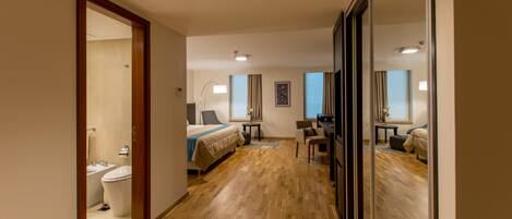A hotel room with a wooden floor, a bed, a desk, a chair, a mirror, and a bathroom with a toilet and sink.