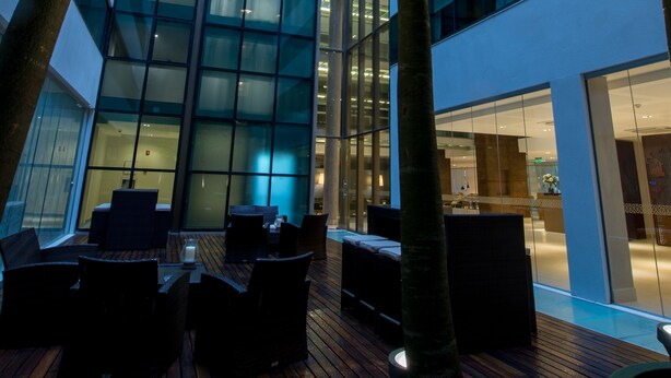 An outdoor seating area with wooden flooring and modern furniture, surrounded by trees and a glass building facade. - Caseros 248 Hotel (Córdoba)