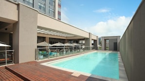 A modern hotel pool area with a wooden deck, lounge chairs, and umbrellas. - Sandton Skye Suite 604 (Sandton)