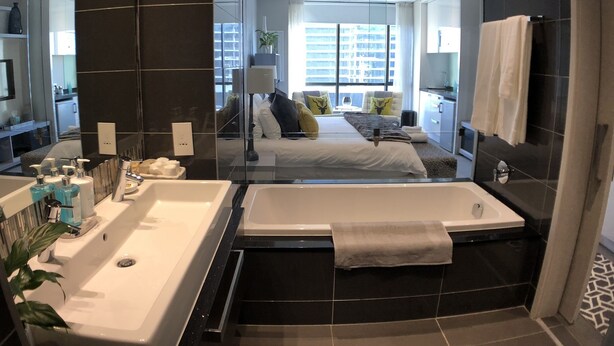 A modern bathroom with a large bathtub, a separate shower area, and a glass-enclosed bedroom with a bed and a view of the city. - Sandton Skye Suite 604 (Sandton)