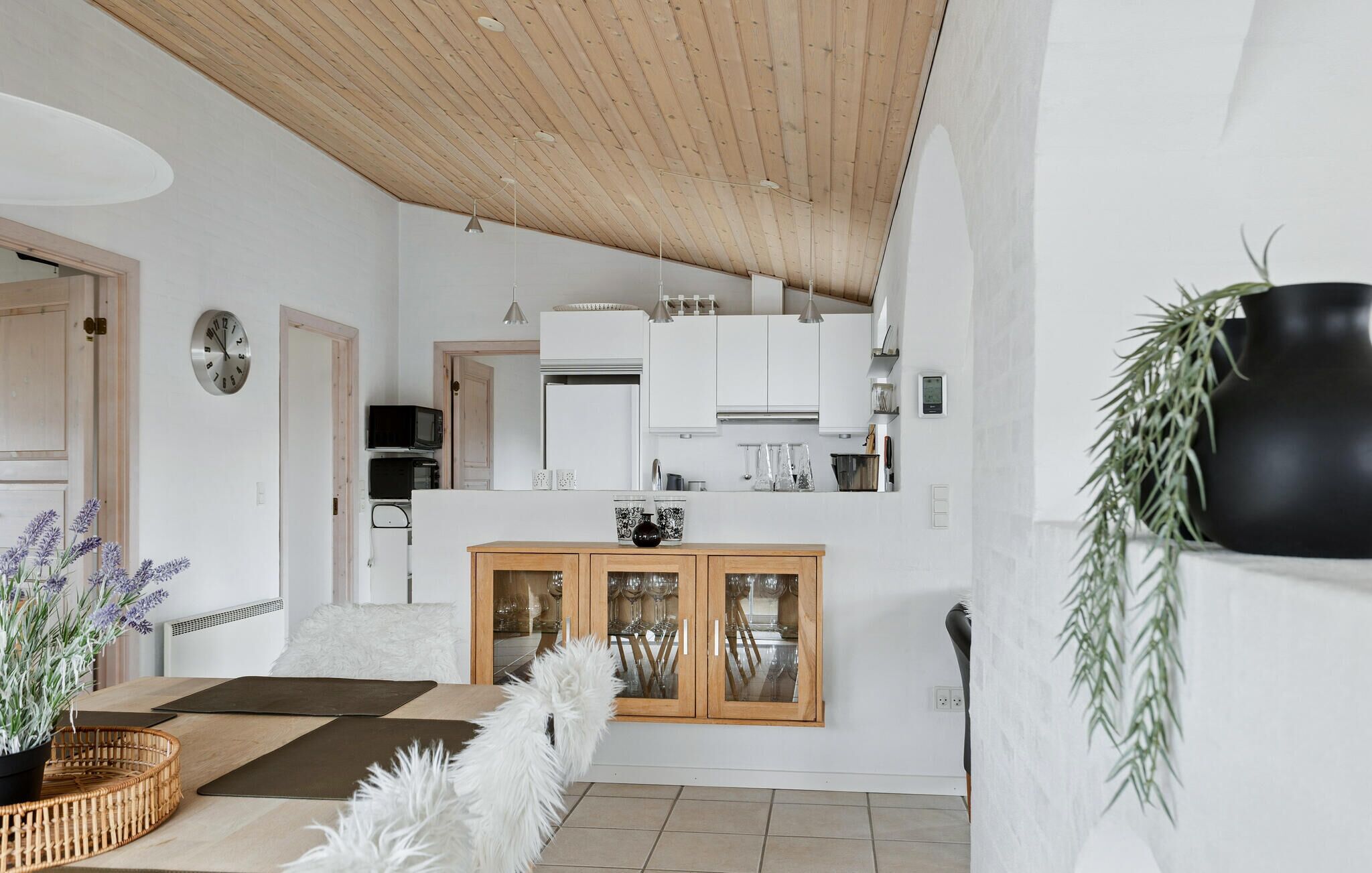 Une cuisine moderne avec des meubles blancs, un plafond en bois et un coin repas agrémenté d’un tapis blanc et moelleux et d’une plante.