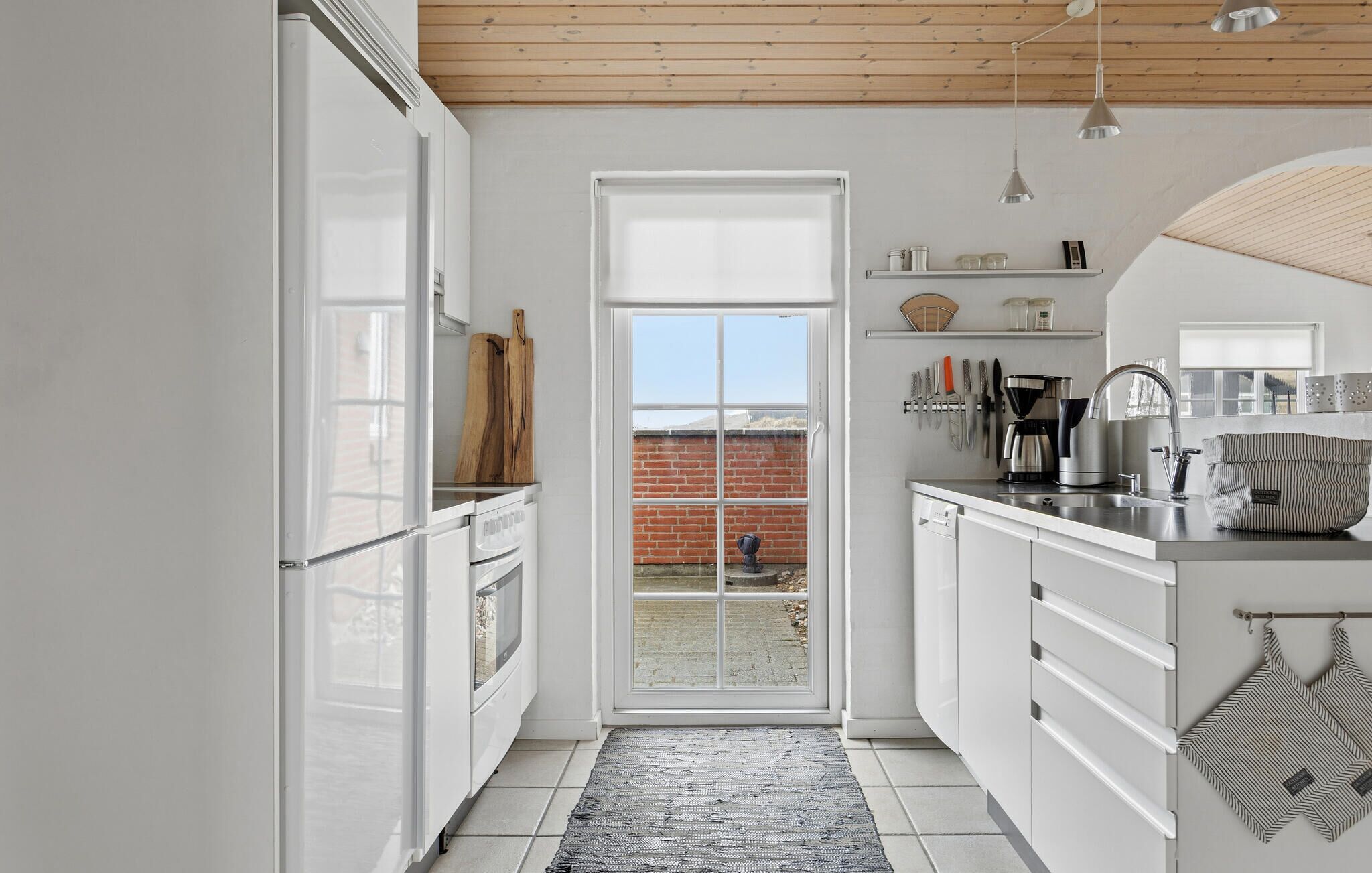 Une cuisine étroite avec des meubles blancs, un plafond en bois et une vue sur un mur de briques, visible à travers une porte vitrée.