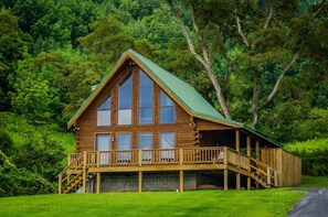 Une cabane en rondins avec un toit végétalisé, de grandes fenêtres et une terrasse en bois avec un escalier menant à un porche abrité.