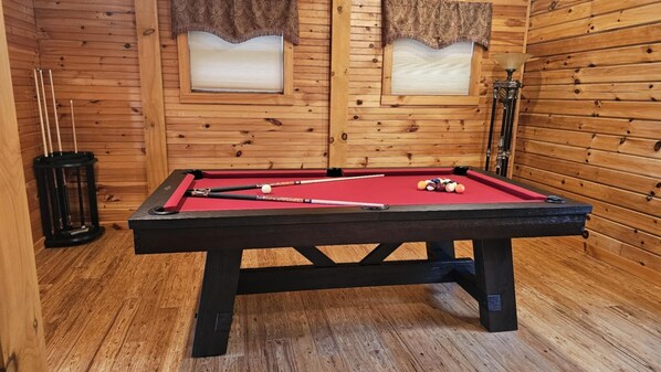 Une table de billard dans une pièce en bois, avec deux fenêtres et une lampe.