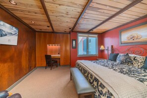 A bedroom with a wooden ceiling, a desk, a chair, a bed with patterned bedding, a bench, and a window with a view of a building.