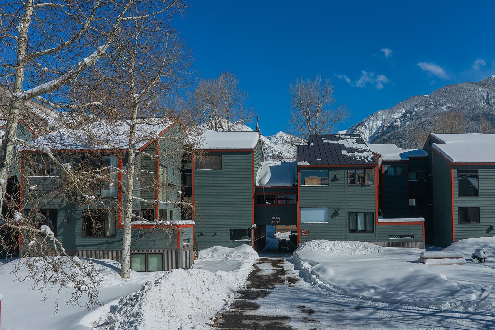 A row of modern, multi-story buildings with a central courtyard, surrounded by snow-covered ground and bare trees, with mountains in the background.