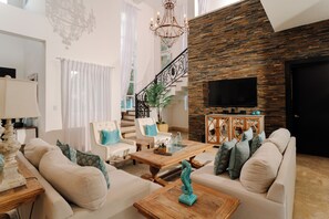 A spacious living room with a staircase, a chandelier, and a flat-screen TV mounted on a wooden wall. - Lifestyle Holidays Villas (Puerto Plata)