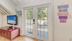 A room with a TV on a wooden cabinet, a door with a view of the outdoors, and a sign on the wall. - Crow's Nest Spinnakers Cottages: 250 Steps to Beach! Pool! (Holmes Beach)