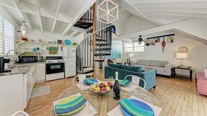 A spacious living area with a kitchen, a dining table with colorful placemats, a blue sofa, and a staircase leading to an upper level. - Crow's Nest Spinnakers Cottages: 250 Steps to Beach! Pool! (Holmes Beach)