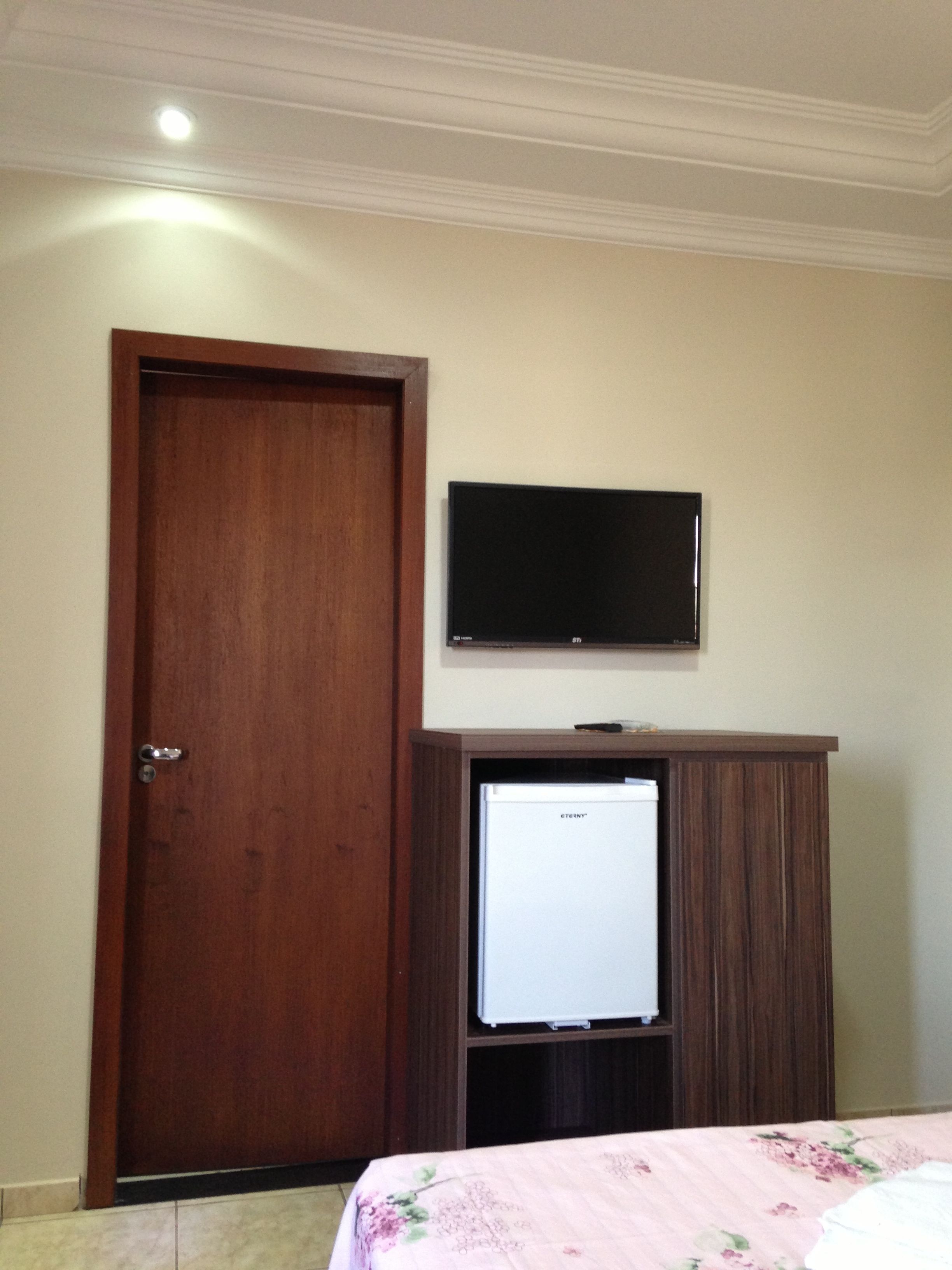 A room with a wooden door, a television mounted on a wooden cabinet, and a bed with a floral bedspread.