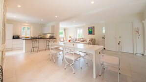A spacious kitchen with a central island, white cabinetry, and a dining area with a white table and metal chairs. - Villa Saint-Tropez (Gassin)
