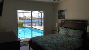 Quarto com cama, mesa de cabeceira, luminária, ventilador de teto e uma grande janela com vista para uma piscina.