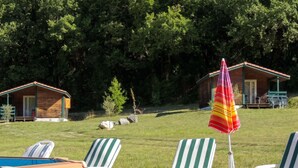 Un área de piscina con sillones de descanso y un paraguas colorido, rodeada de cabañas de madera y un campo cubierto de césped.