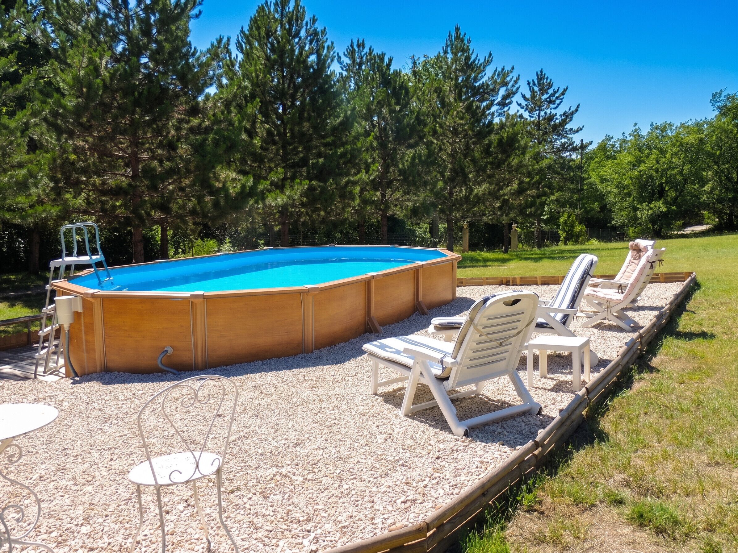Une piscine extérieure dotée d’une terrasse en bois, entourée d’arbres et aménagée avec des chaises longues et une petite table.