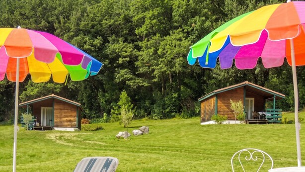 Una piscina con una tumbona y una pequeña mesa, dos cabañas de madera con mobiliario exterior y un paraguas colorido.