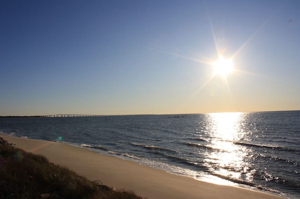 A beach with a clear sky, a sun reflecting on the water, and a distant bridge.