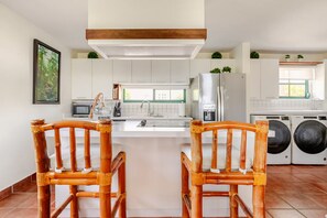 A modern kitchen with white cabinets, a stainless steel refrigerator, a built-in oven, and a washing machine. There are two wooden chairs with woven seats.