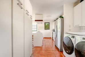 A narrow kitchen with a washing machine, a refrigerator, and a window with a view of greenery.