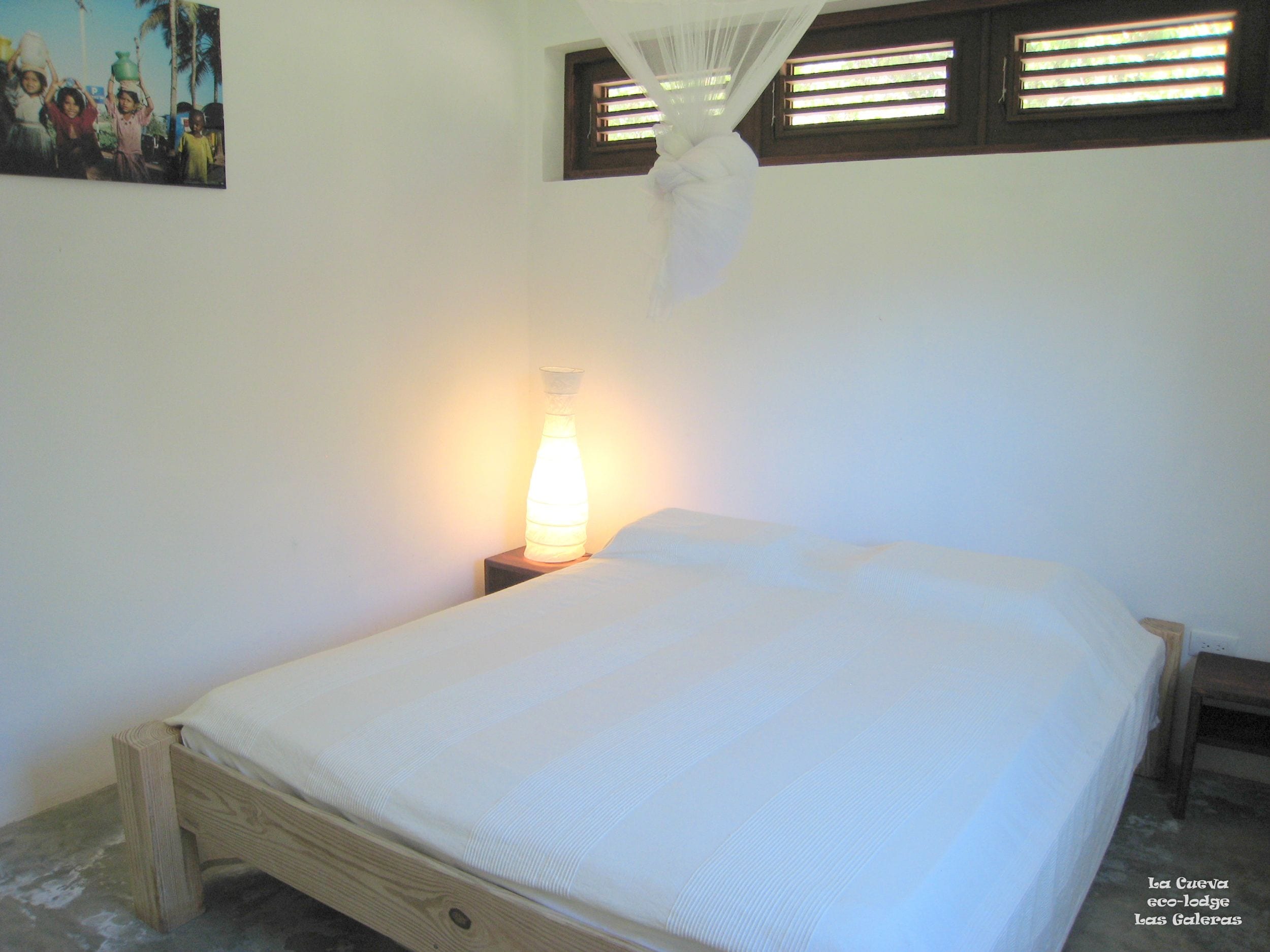 Una cama individual con colchón blanco y estructura de madera, una mesita de noche con lámpara, y una pared blanca lisa.