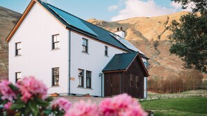 Une maison blanche avec un toit sombre et une grange en bois, entourée d’un paysage verdoyant et de collines.