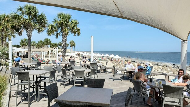An outdoor seating area with tables and chairs, overlooking a beach with umbrellas and people.