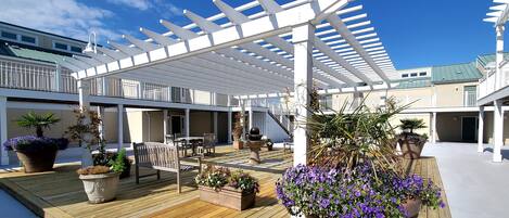 A covered outdoor deck with wooden flooring, white pergola, and potted plants.