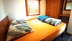 A double bed with a wooden headboard, two pillows with a leaf pattern, a wooden wardrobe, and a window with blinds.
